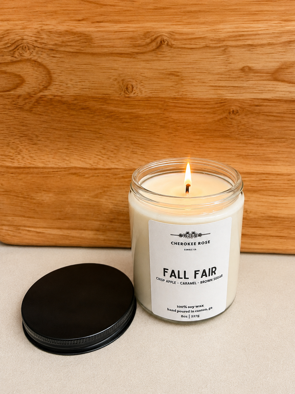 A lit candle in a clear glass jar with a white label reading 'Cherokee Rose Candle Company' and 'Fall Fair,' placed on a light surface with a black lid beside it and a wooden background.