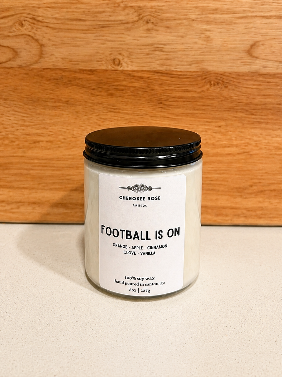 A candle jar labeled 'FOOTBALL IS ON' by Cherokee Rose, with white wax, a black lid, and a white label, set against a light wood background and light surface.

Details