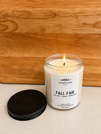 A lit candle in a clear glass jar with a white label reading 'Cherokee Rose Candle Company' and 'Fall Fair,' placed on a light surface with a black lid beside it and a wooden background.